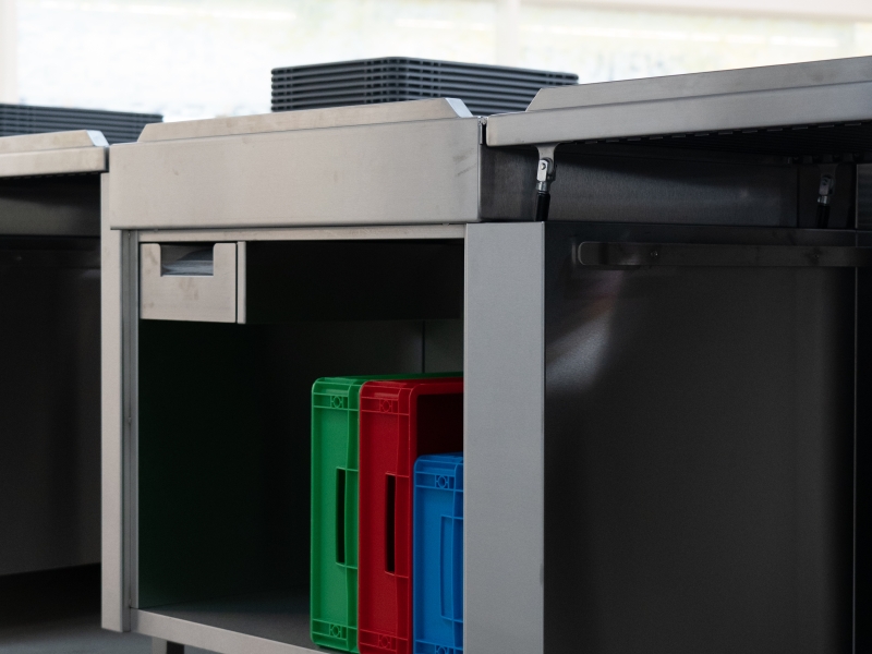 Edelstahl-Möbel mit individuell höhenverstellbaren Füßen, offenem Arbeitsschrank für Kisten und Arbeitsgüter, integrierter Sonder-Lade für Sicherheitsbeutel und Durchgangsklappe mit Gasdruckdämpfer