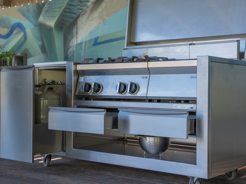 Foto zur Präsentation der Funktionen des Outdoormöbels: Links = Schwerlastauszug zum Verstauen der Gasflasche; Mittig = 2x Lade für Kochgeschirr- und Besteck; Oben = Klappbare CNS-Haube für wetterfestes Verstauen der Kochgeräte;