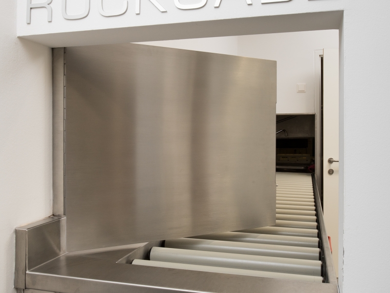 Geschirr-Rückgabe in Sonder-Ausführung: Geschirr rollt durch Gefälle und Rollen direkt zum Haubenspüler; Durchführung durch zwei Mauern; verschließbar durch CNS-Tür