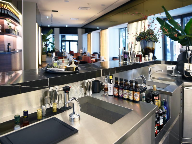 Aufnahme aus Sicht des Barkeepers mit Sicht auf die Edelstahl-Verbauten der Bar und den Gästebereich im Hintergrund